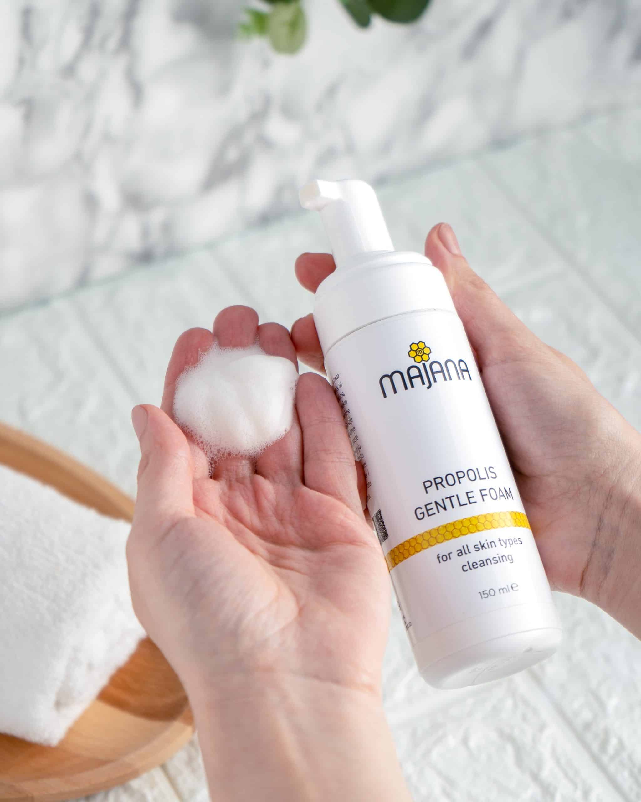 En person håller en vit flaska Majana Propolis Gentle Foam i ena handen och en liten mängd skum i den andra. Flaskan har en gul och svart logotyp med text som indikerar att den är för rengöring av alla hudtyper. I bakgrunden finns en vit handduk på en träbricka och vita kakel.